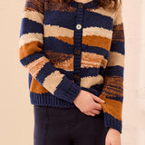 A woman wearing a chunky knit cardigan sweater with horizontal stripes in navy blue, beige, and rust orange, paired with dark pants.