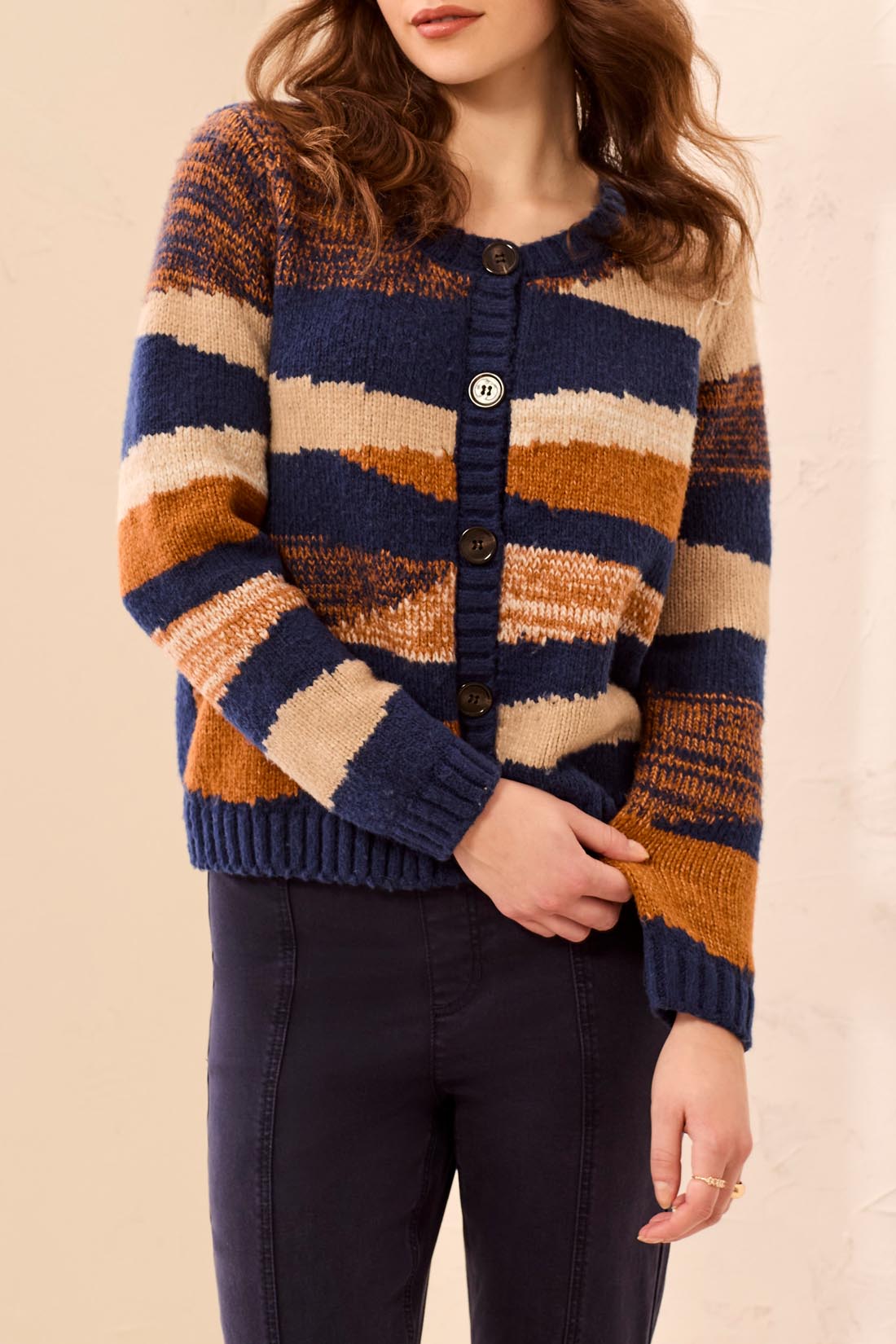 A woman wearing a chunky knit cardigan sweater with horizontal stripes in navy blue, beige, and rust orange, paired with dark pants.