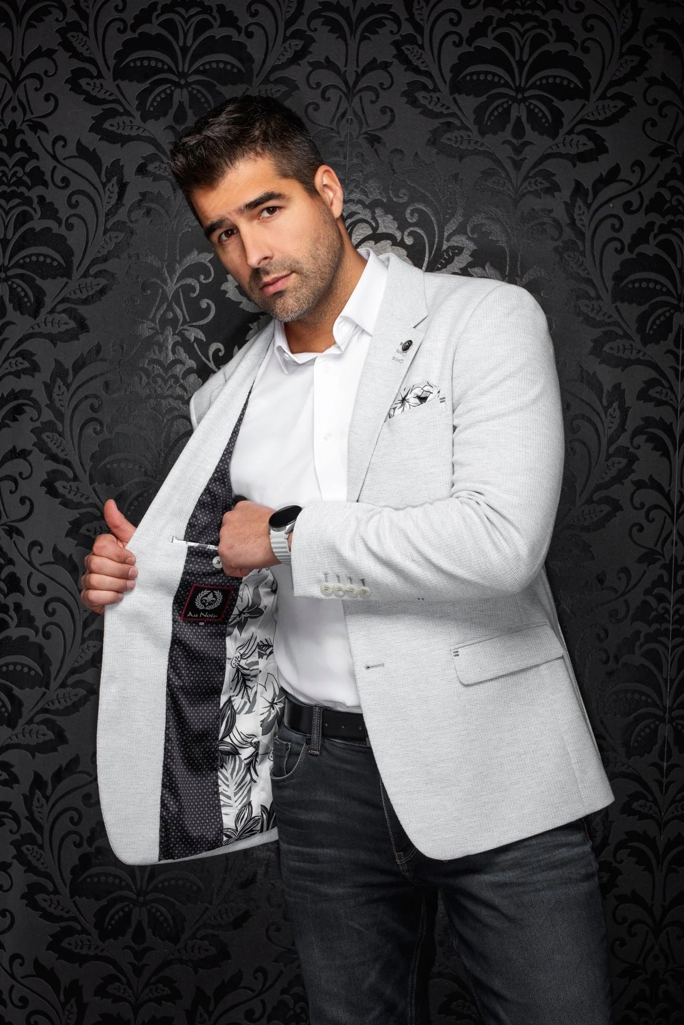 Man in a white blazer over a patterned shirt against a dark, ornate background