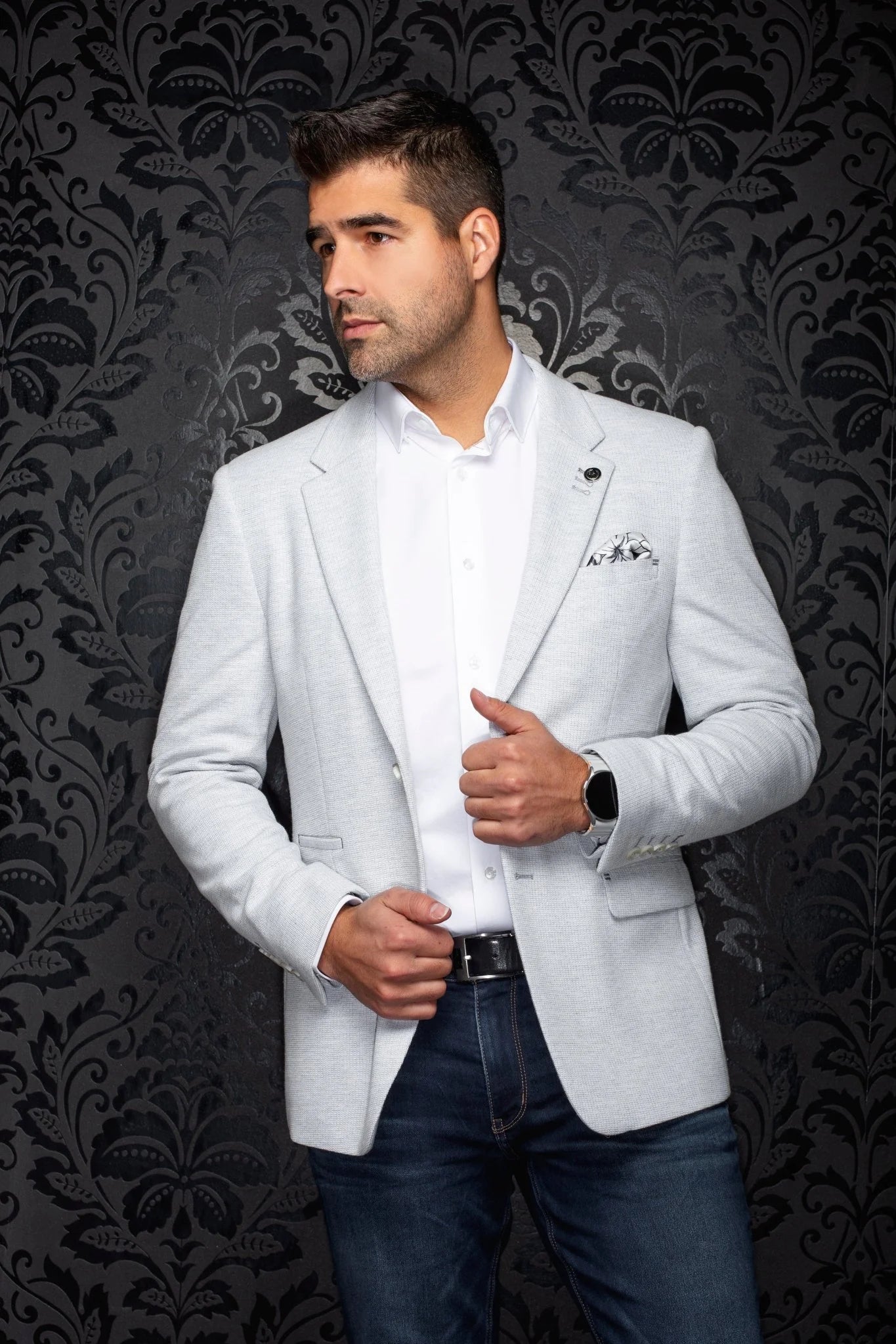 Man wearing a light gray blazer over a white shirt with a dark patterned background